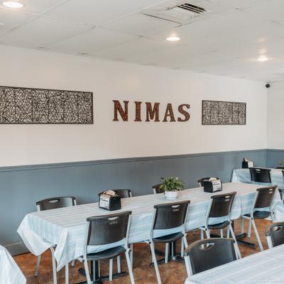 A bright, spacious dining area with blue tablecloths, black chairs, and a wall sign reading Nimas.