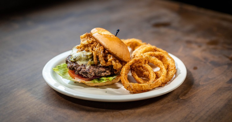 A burger topped with onion rings sits on a plate with sides