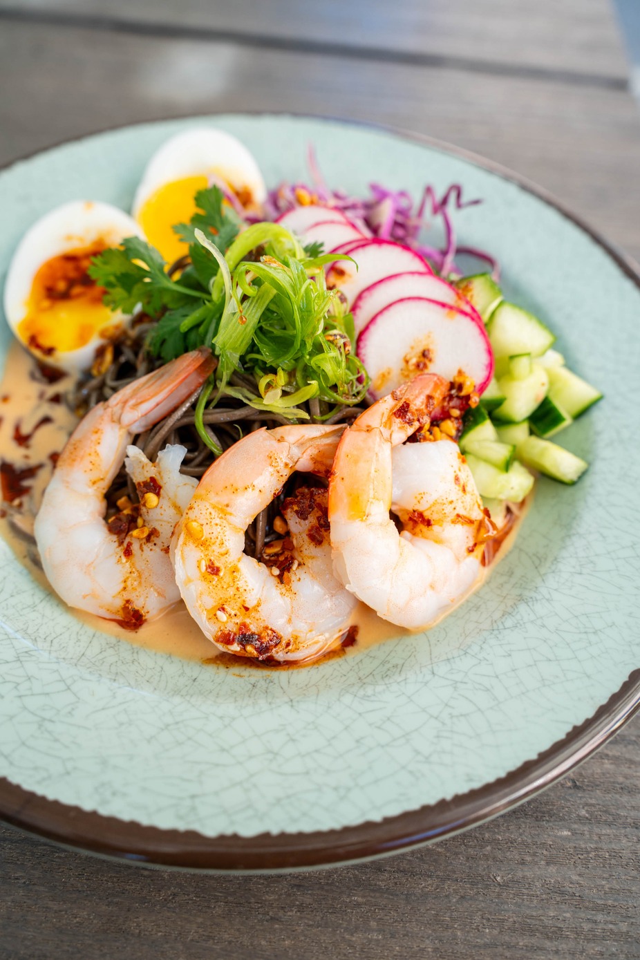 Chilled Shrimp Noodle Bowl- Cold Buckwheat noodles tossed in a creamy miso broth, topped with purple cabbage, cucumber, radish, cilantro, green onion, and finished with chili crunch, a runny egg, and poached shrimp.