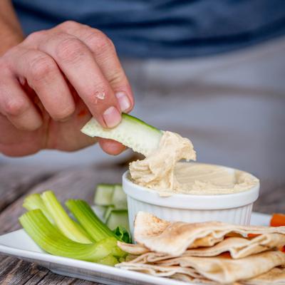 A person dips a celery stick into hummus on the plate with pita and veggies.