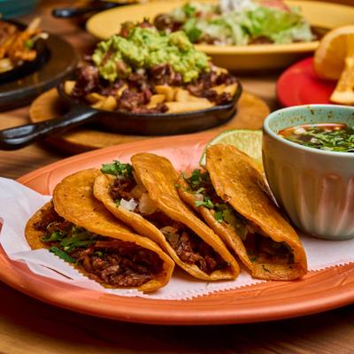 Three birria tacos served with dipping broth and other dishes in the background.