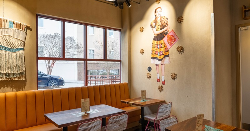 Cozy interior with a mural of a girl on the wall, wooden tables, orange bench and large window.