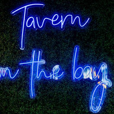 Blue neon sign reading 'Tavern on the bay' against a leafy backdrop.