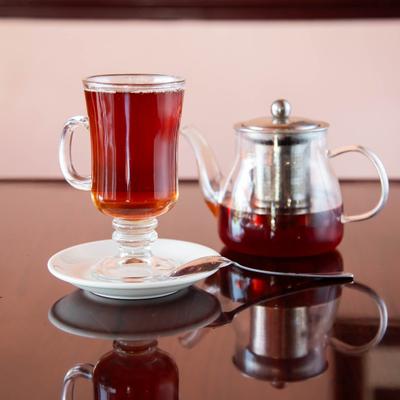 Red tea served in a tall glass with handle and a glass teapot on a table.
