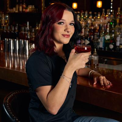 An employee sits at a bar, holding a red cocktail and smiling.