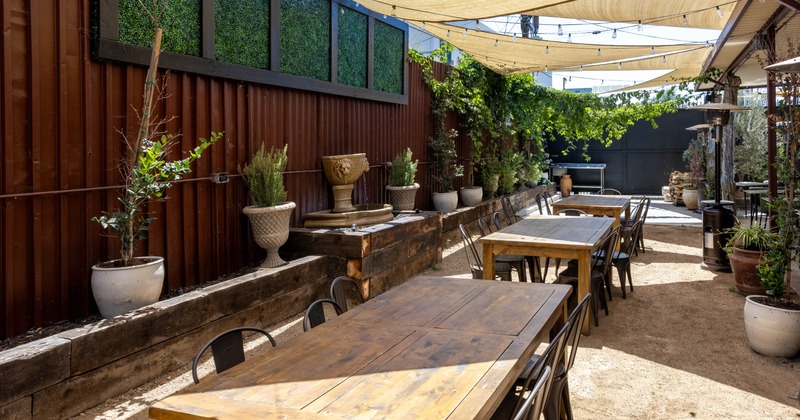 Outdoor dining area with wooden tables, metal chairs, and shade sails overhead