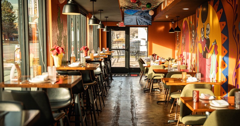 Interior of a brightly lit restaurant with tables, chairs, large windows, murals