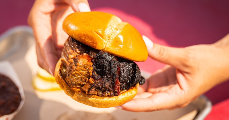 Close-up of hands holding a brisket sandwich with a shiny bun and juicy, charred meat