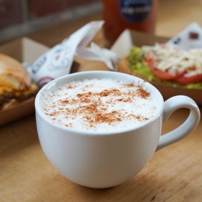 A cup of frothy cappuccino sprinkled with cinnamon sits on a table with food.