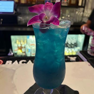 A glass of Blue Hawaiian  with a pink orchid flower, sits on a bar counter.