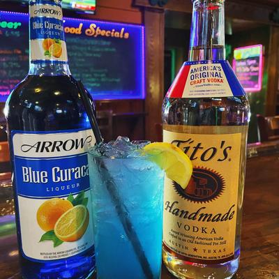 A blue cocktail is garnished with a lemon slice and placed between bottles of liqueur and vodka.