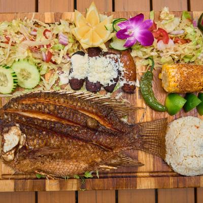 Fried whole tilapia fish served with sides like rice, salad, corn, and avocado.