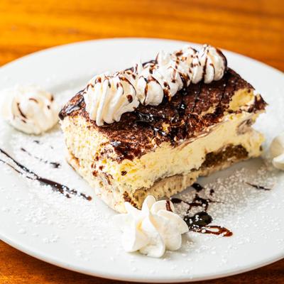 A slice of tiramisu  topped with whipped cream and chocolate drizzle on a white plate.