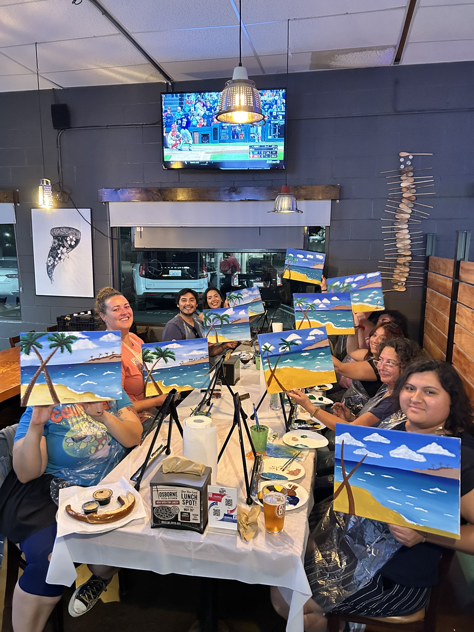 Guests at Osborne Pizza Kitchen display colorful beach paintings during a Paint N Sip event