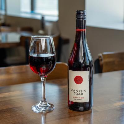 A bottle of red wine and a glass of red wine sit on a wooden table in a softly lit room.