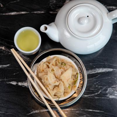 A white teapot and a cup beside a bowl of beef tripe dim sum with chopsticks.
