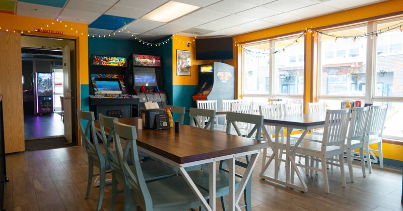 Brightly colored dining area with arcade machines, wooden tables & light blue chairs,