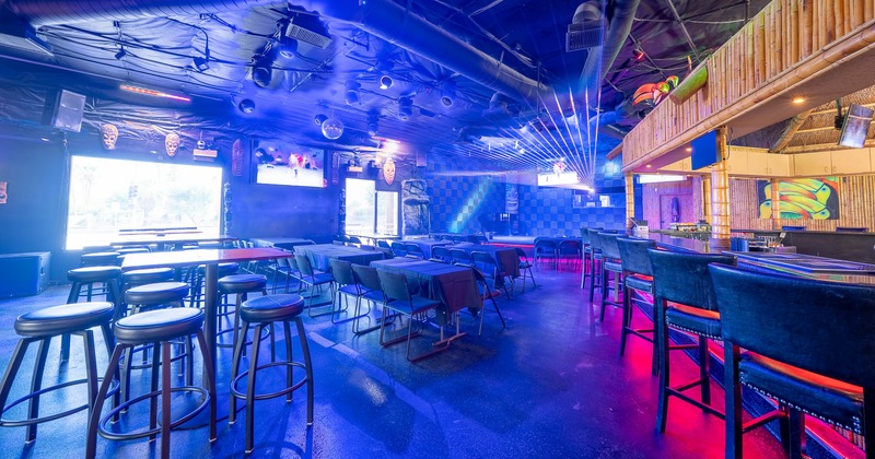 Interior of a colorful bar with blue and pink lighting, tables, stools, and a bar counter.