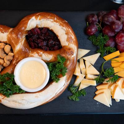 A black platter with a pretzel, cashews, cranberries, cheese dip, grapes, and sliced cheeses.