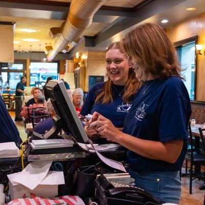 Two employees t employees work together at a point-of-sale station inside a dining area.