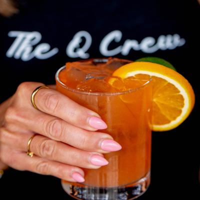 Manicured hand wearing golden rings holding an orange cocktail on the rocks with citrus wheels.