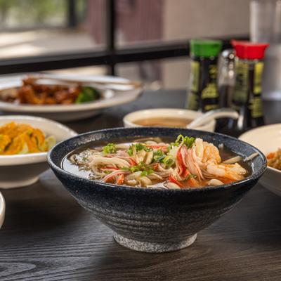 A bowl of Seafood Udon soup sits on a dark table, surrounded by various other dishes.