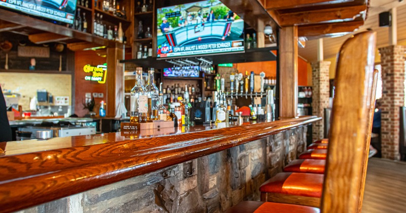 Interior of a bar - a wooden counter, orange-cushioned stools and flat-screen TVs displaying sports