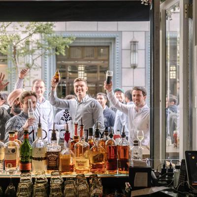 A group of smiling people stands outside a bar window, raising drinks in a cheerful toast.