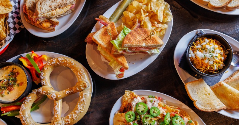 A table laden with sandwiches, pretzel with dip, nachos, and a bowl of chili