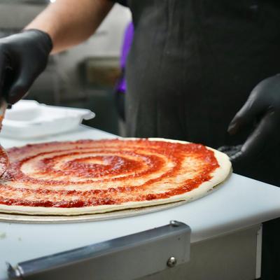 A person in black gloves spreading tomato sauce on pizza dough.