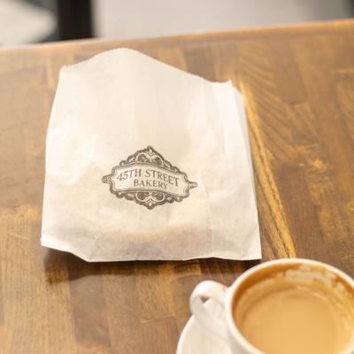 A small paper bag sits next to a cup of coffee on a wooden table.