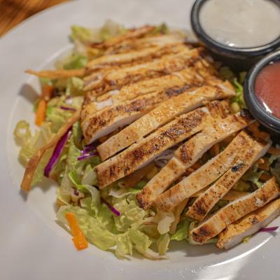 Grilled chicken salad with lettuce, carrots, and dressing cups on a white plate.