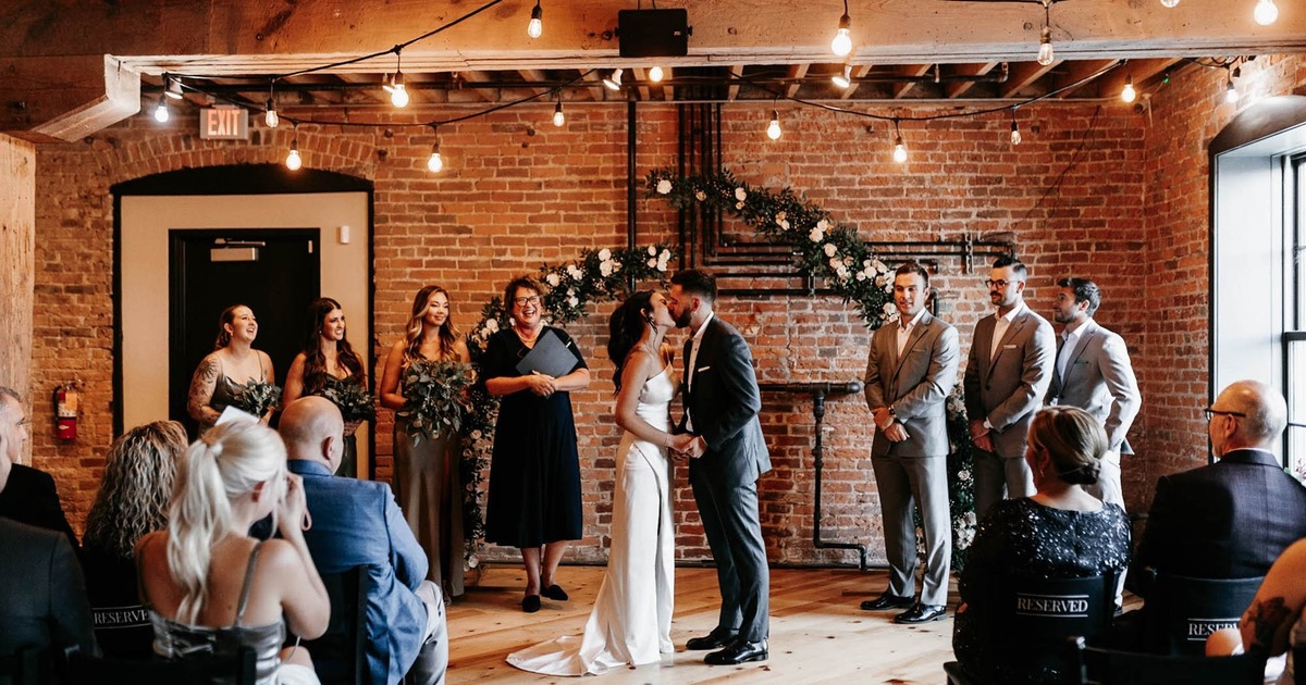 Spacious rustic room with wooden beams and floors, brick walls, windows, tables, and chairs
