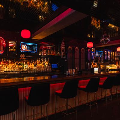 Dimly lit bar counter with red lighting, stools, and illuminated shelves of liquor.