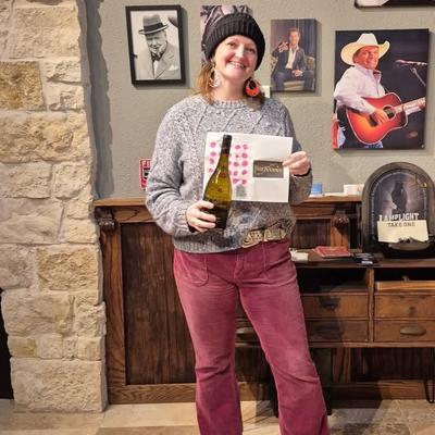 Person standing in front of the wall with framed photos, holding a wine bottle.