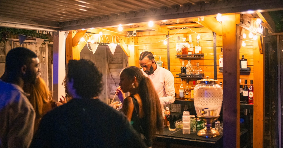 Outdoor bar scene at night, with warm lighting and several people engaging with a bartender