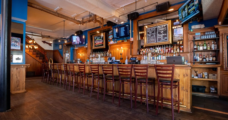 Interior, long wooden counter lined with tall chairs, shelves filled with liquor bottles
