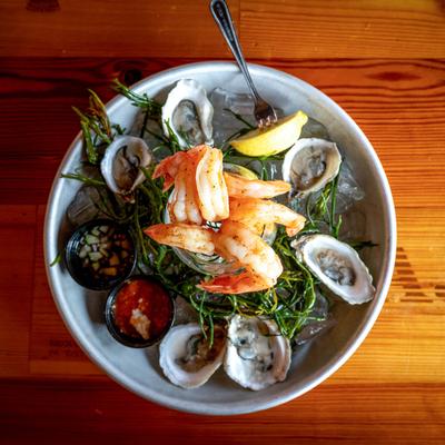 A glass filled with ice and shrimp is placed on a plate of oysters with sauces and lemon.