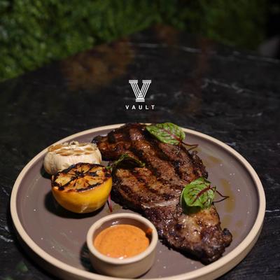 A grilled steak served with grilled lemon half, garlic, and a side sauce.
