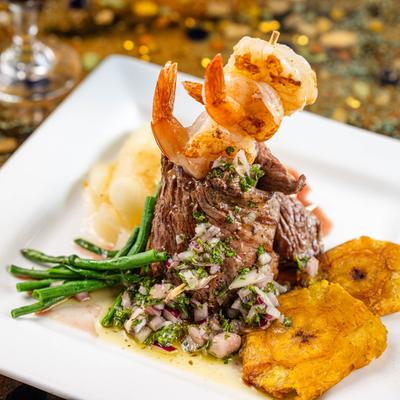 A grilled steak topped with shrimp and chimichurri, served with fried plantains.