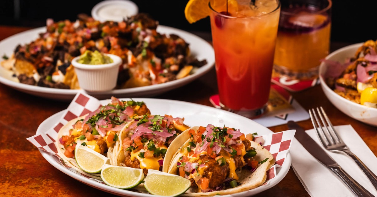 A plate with fish tacos on a wooden table with nachos and a glass of Tequila Sunrise