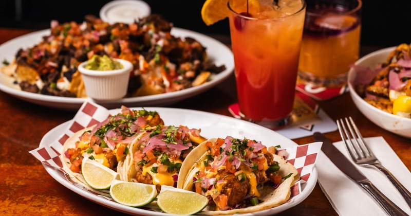 A plate with fish tacos on a wooden table with nachos and a glass of Tequila Sunrise