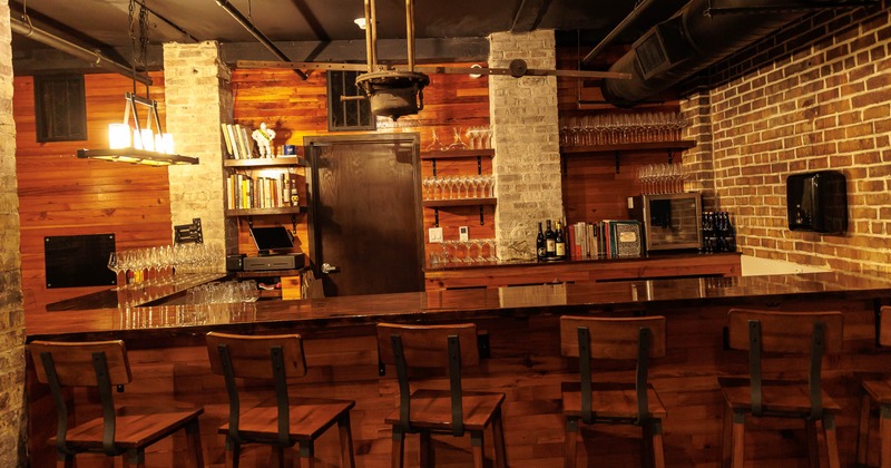 A bar interior - a polished wooden counter, tall chairs, exposed brick, wood paneling, glassware.