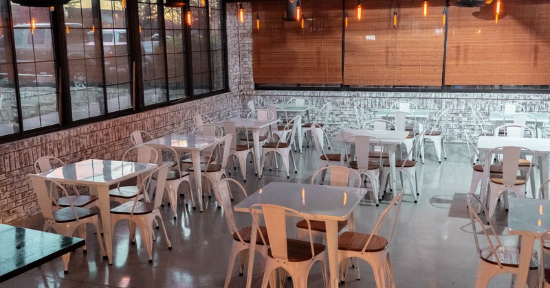 An indoor restaurant seating area with white tables and chairs, pendant lights, and large windows