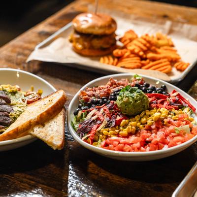 Table spread featuring burrito bowl, burger with fries and a plate of pasta with a side of toast.