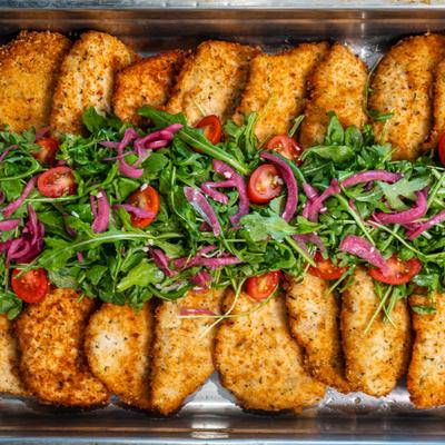 Breaded chicken cutlets topped with arugula and cherry tomatoes in a catering tray.