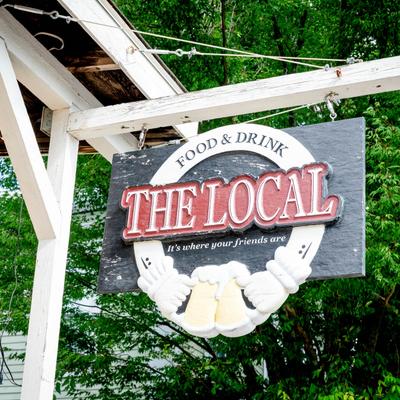 The Local restaurant sign hanging outside with trees in the background.