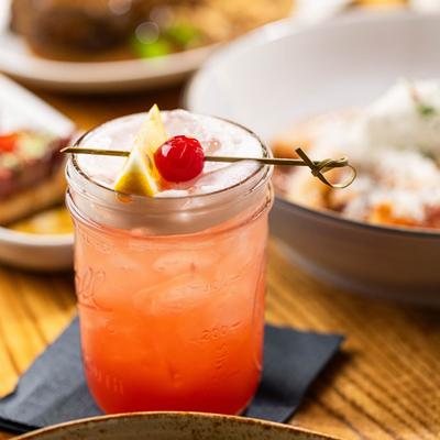Pink cocktail in a mason jar, garnished with a lemon wedge and a cherry.