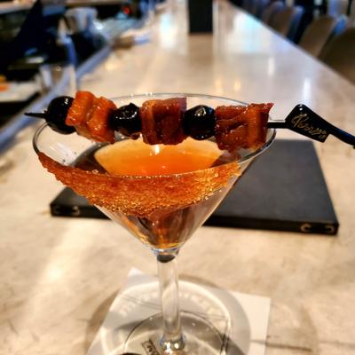Cocktail in martini glass garnished with a skewer of pork chop bites and cherries.