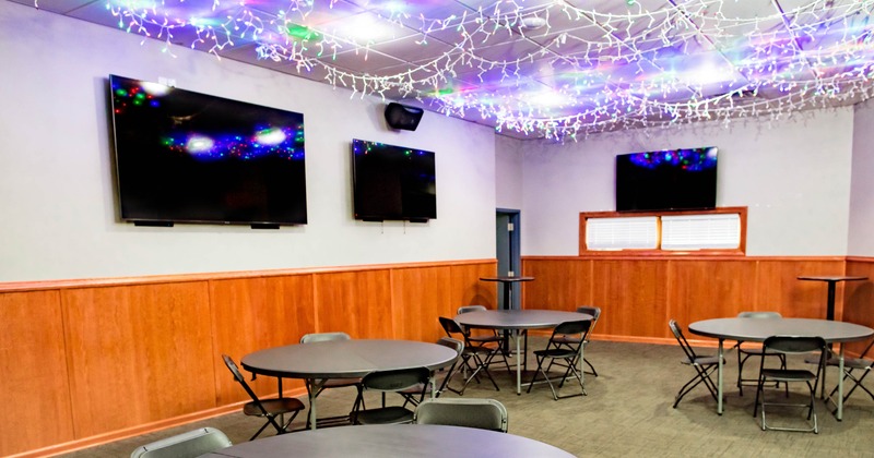 Room with round tables, folding chairs, mounted TVs, and ceiling string lights